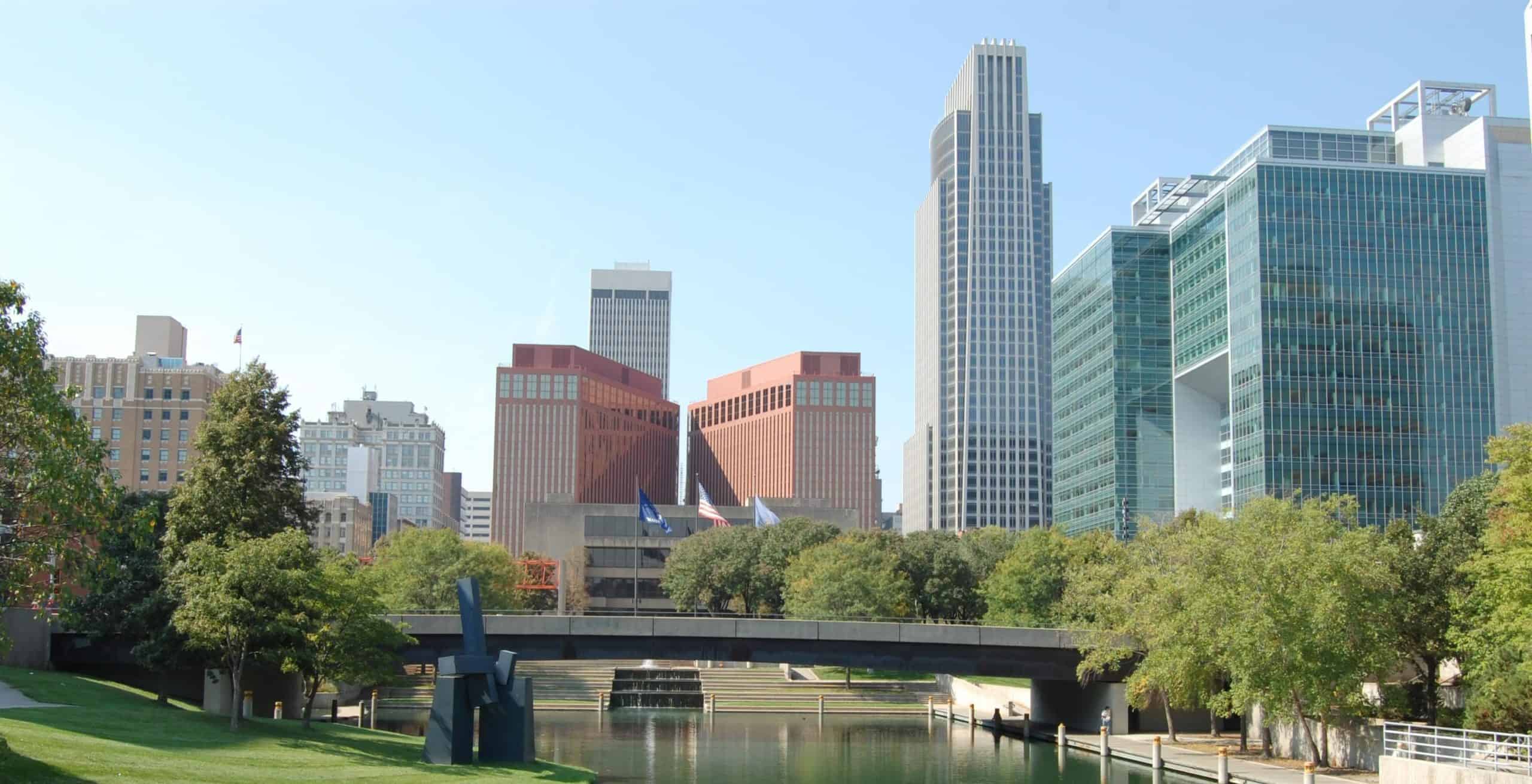 Omaha skyline: modern and historic buildings, high-rises, trees by a waterway under a clear blue sky.